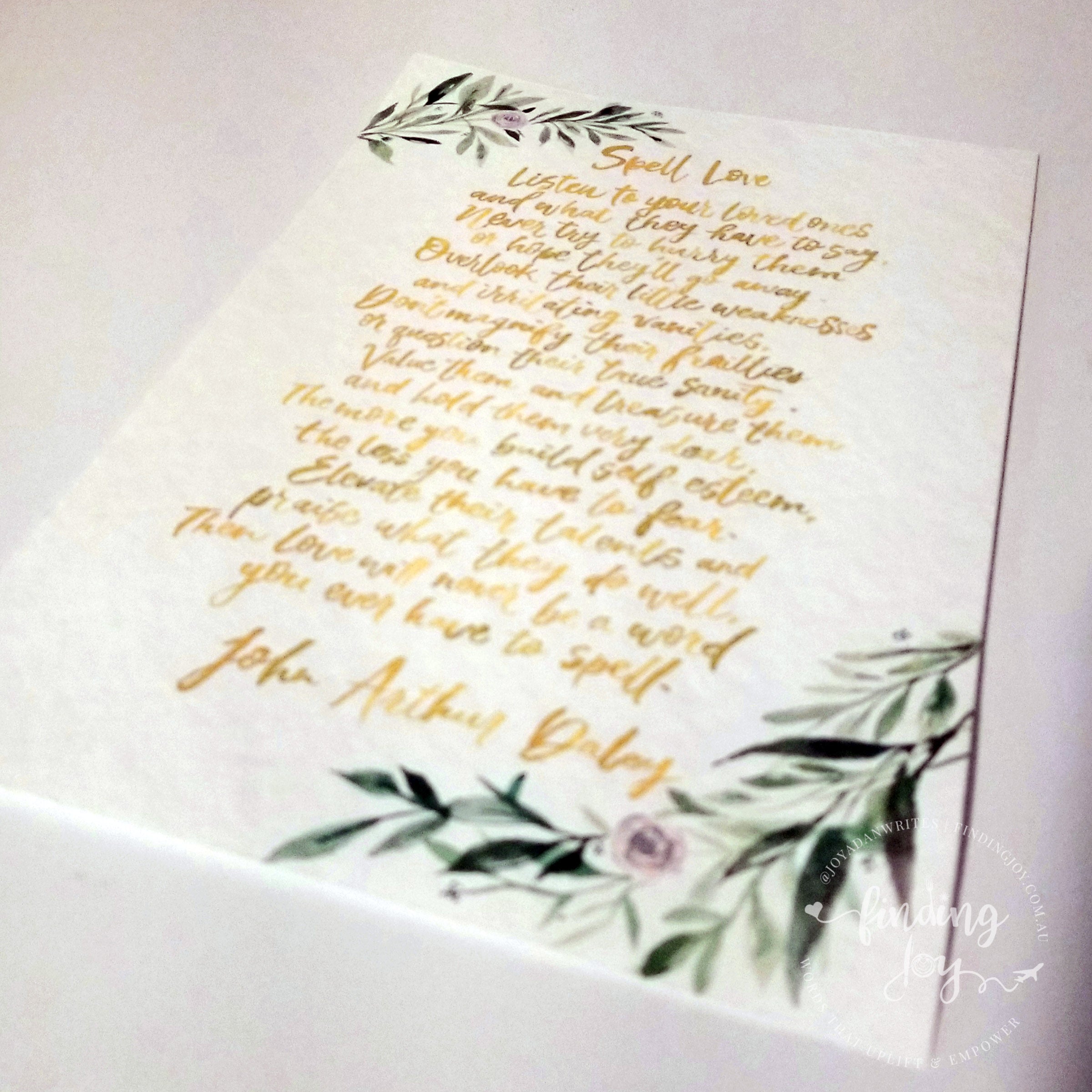 A square white card featuring gold foil calligraphy of 'Spell Love' from John Anthony Duboy's 'Little Awakening' poem, decorated with green watercolor leaves and a small pink rose at the bottom, shown on a white background.