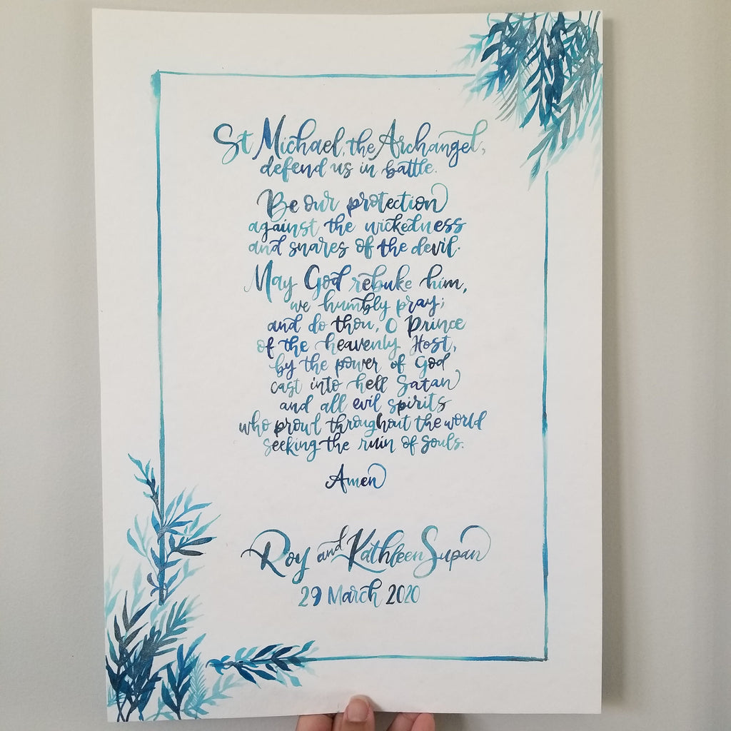 A vertical white printed card with blue watercolor botanical corner details and blue script text, held against a light neutral background. The card appears to be a personalized religious print or invitation featuring St Michael the Archangel and the names and date of a couple.