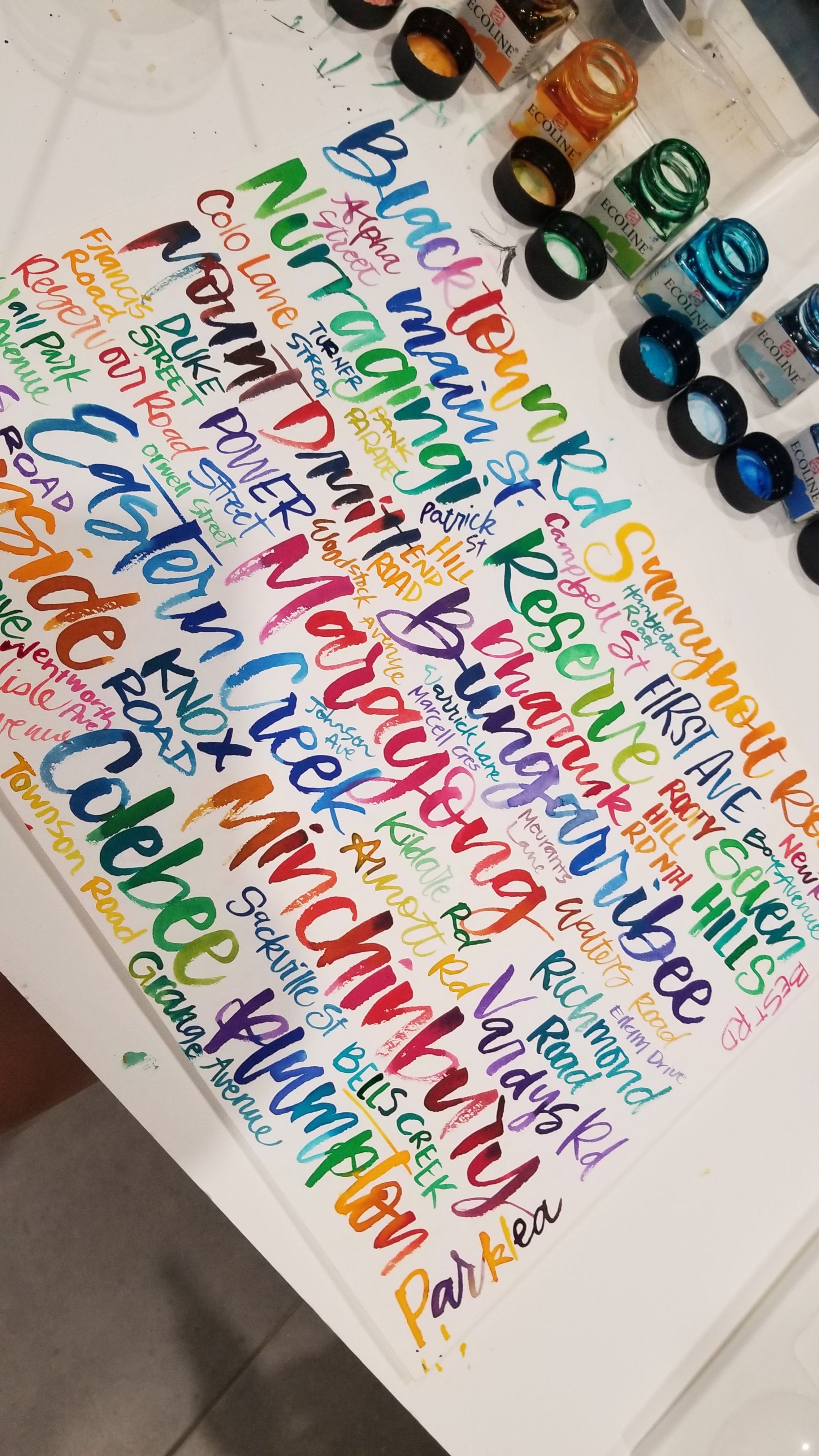 A close-up of a white canvas covered with colorful hand-lettered place names and words, with paint tubes arranged along the top edge.