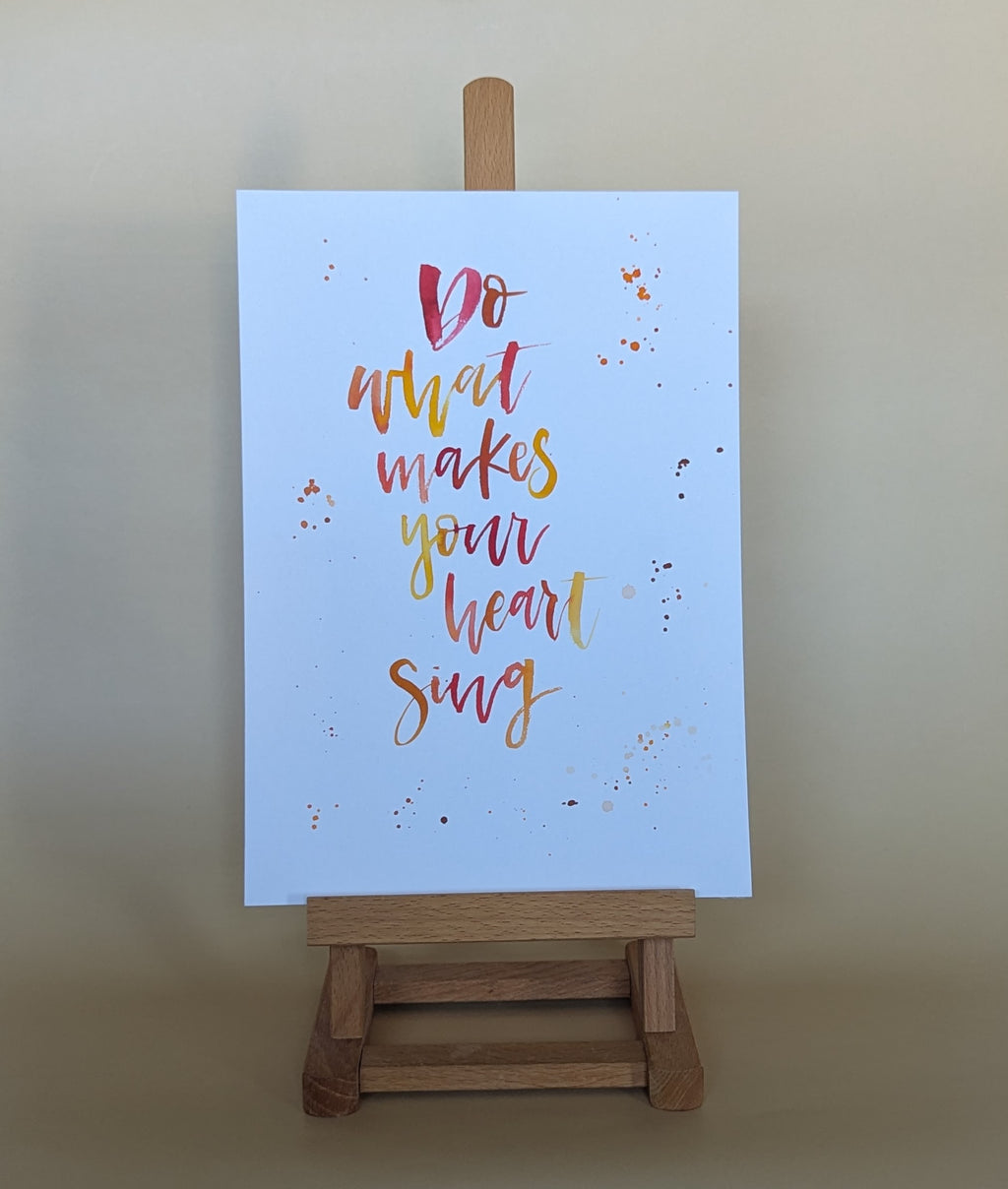 A vertical art print with the hand-lettered quote 'Do what makes your heart sing' in red, orange, and yellow on a light blue background, displayed on a small wooden easel against a warm beige wall.