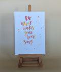 A vertical art print with the hand-lettered quote 'Do what makes your heart sing' in red, orange, and yellow on a light blue background, displayed on a small wooden easel against a warm beige wall.