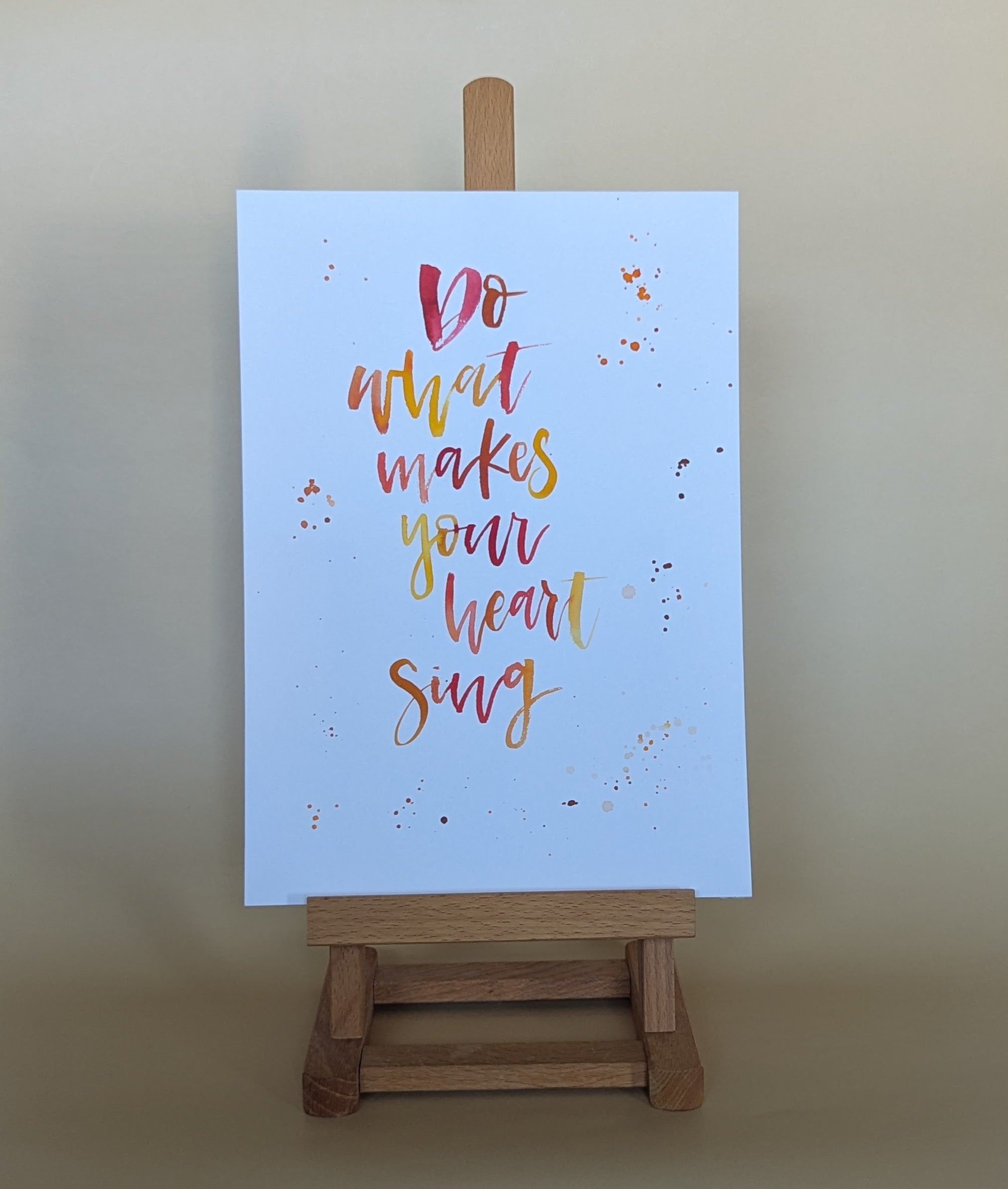 A vertical art print with the hand-lettered quote 'Do what makes your heart sing' in red, orange, and yellow on a light blue background, displayed on a small wooden easel against a warm beige wall.