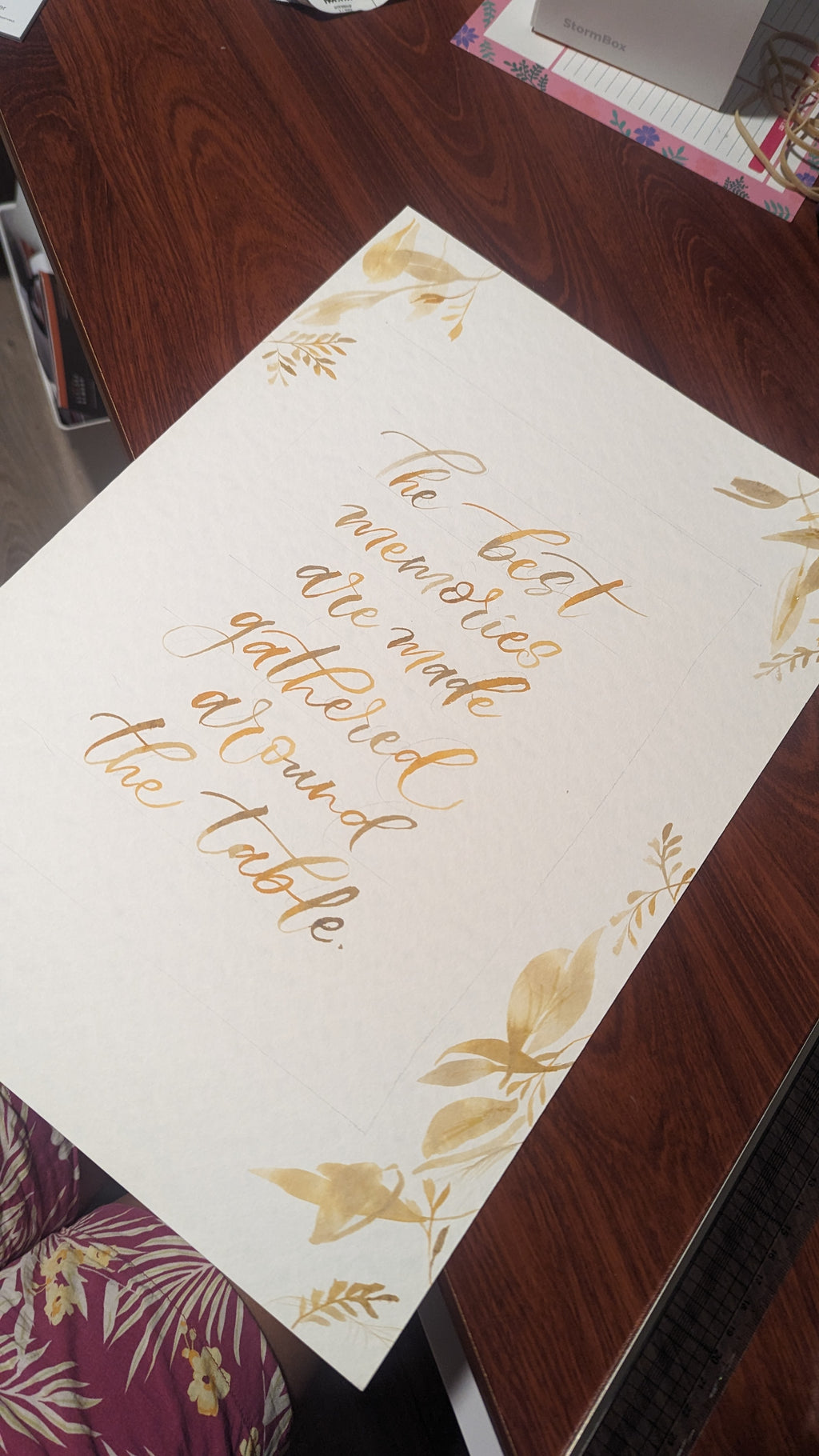 A square white card with elegant gold foil calligraphy reading 'the best memories are made around the table' and gold floral corner accents, photographed on a dark wooden tabletop.