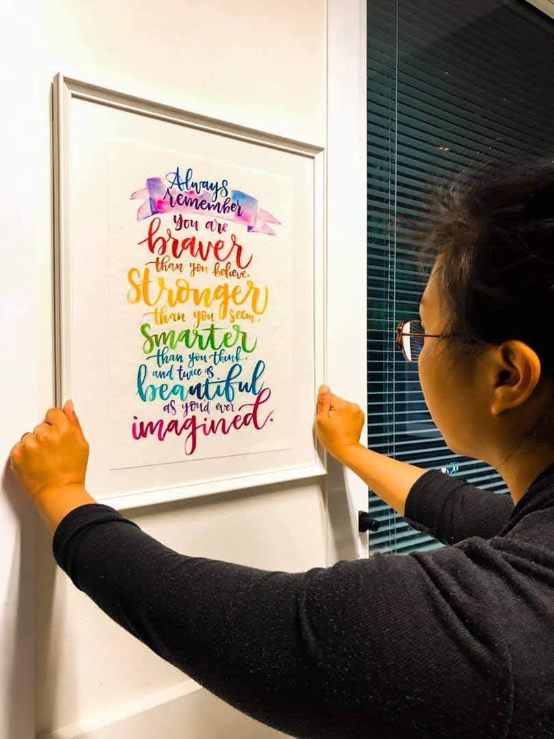 A person holding a white-framed inspirational art print with colorful hand-lettered text, mounted on a white wall next to a window with dark blinds.