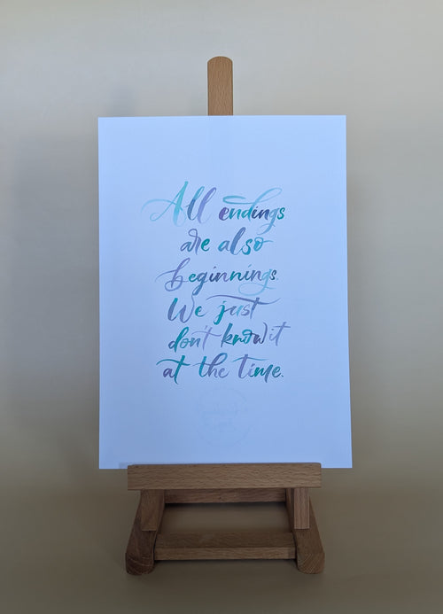 A vertical art print with a quote in blue and teal cursive lettering on a white sheet of paper, displayed on a small wooden easel against a neutral beige background.