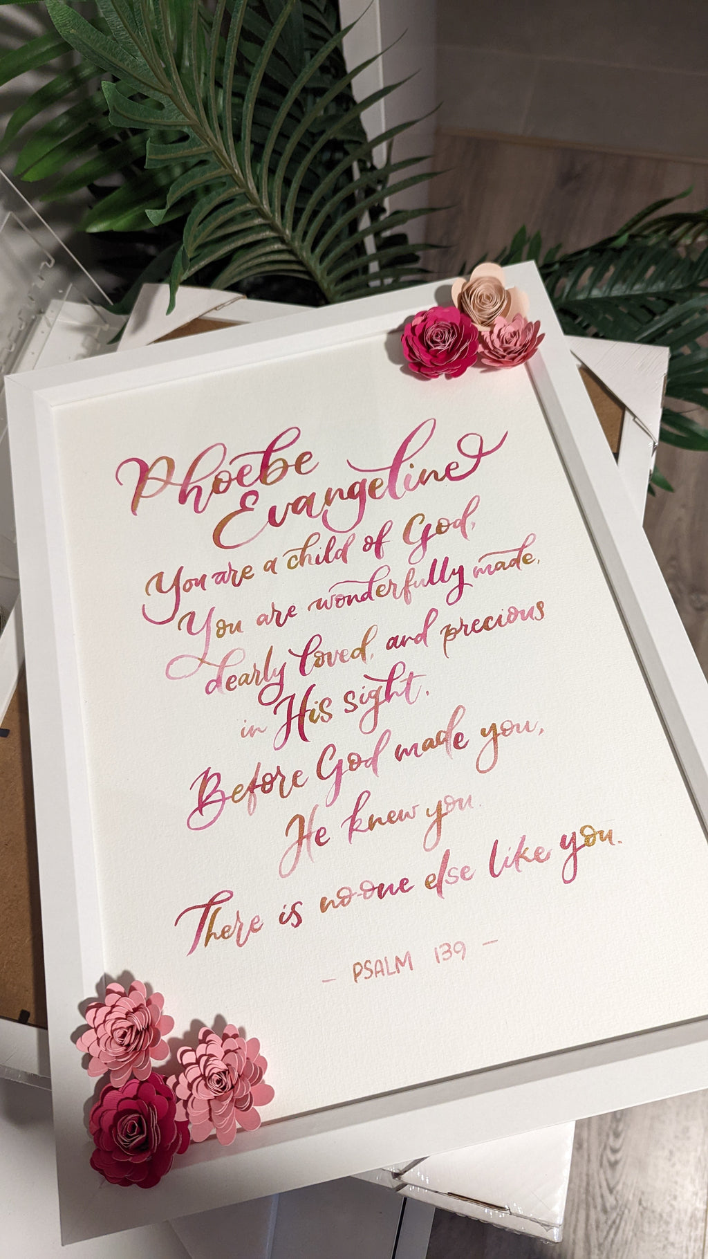 A framed typographic wall art print featuring a Bible verse in pink and gold calligraphy on a white background, styled with pink and beige flowers in the corners and leaning against a surface with green leaves in the background.