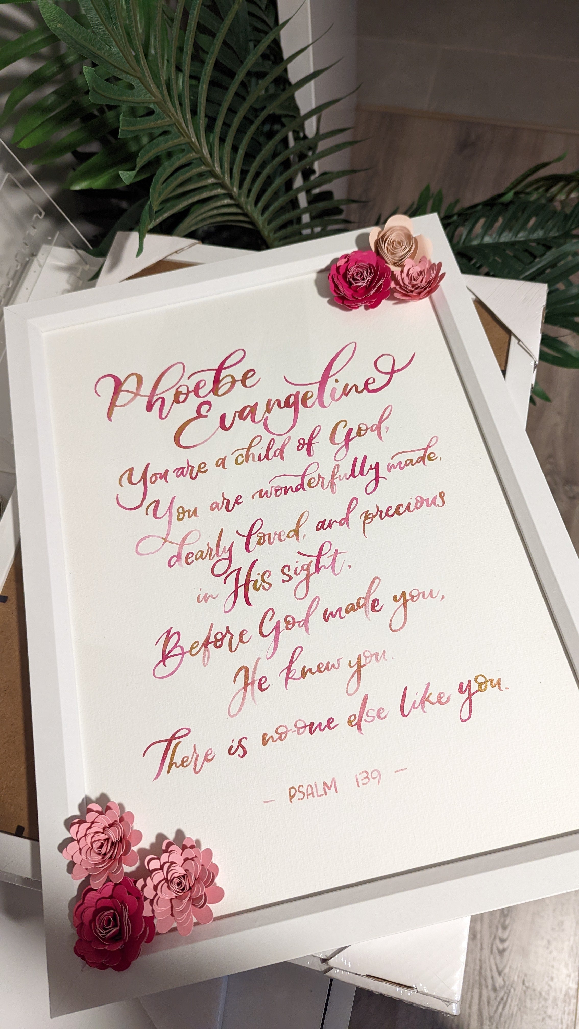 A framed typographic wall art print featuring a Bible verse in pink and gold calligraphy on a white background, styled with pink and beige flowers in the corners and leaning against a surface with green leaves in the background.