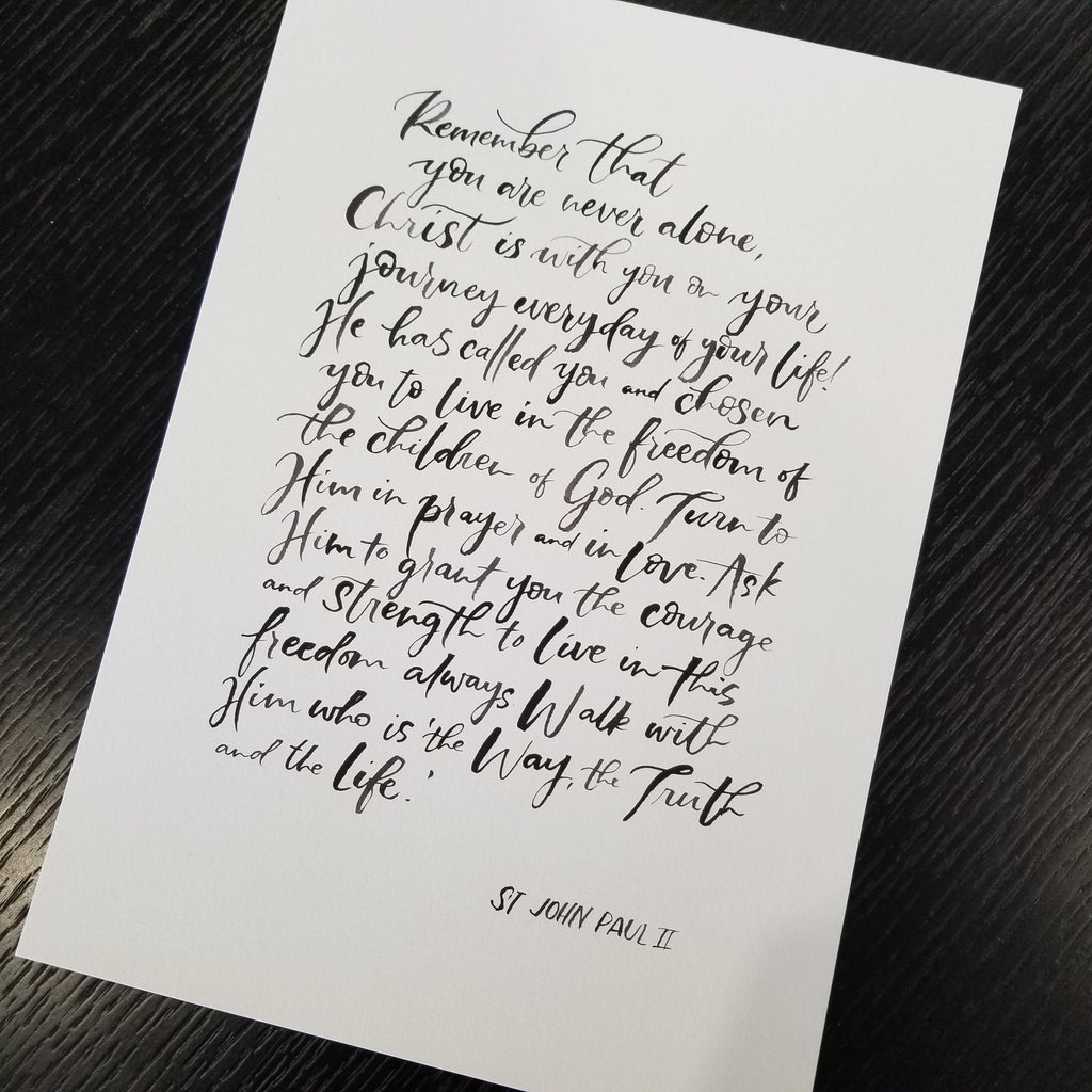 A white art print featuring elegant black calligraphy of a St. John Paul II religious quote, displayed at an angle on a dark wood background.