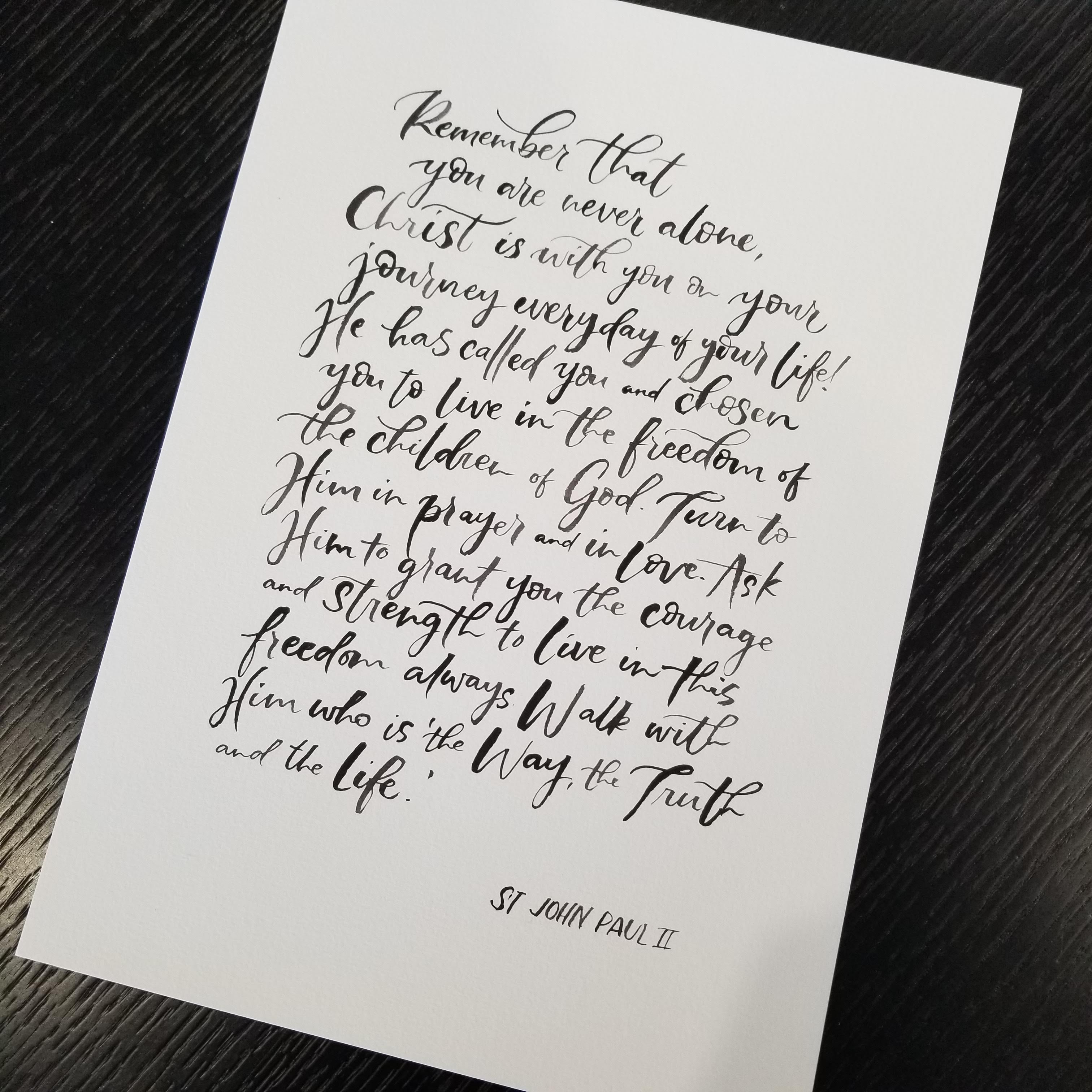 A white art print featuring elegant black calligraphy of a St. John Paul II religious quote, displayed at an angle on a dark wood background.