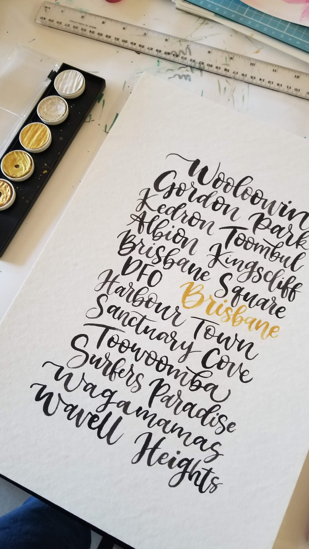 A close-up of a hand-lettered art print featuring various Brisbane suburbs in elegant black and gold calligraphy, displayed on a white paper sheet with a paint palette and tools in the background.