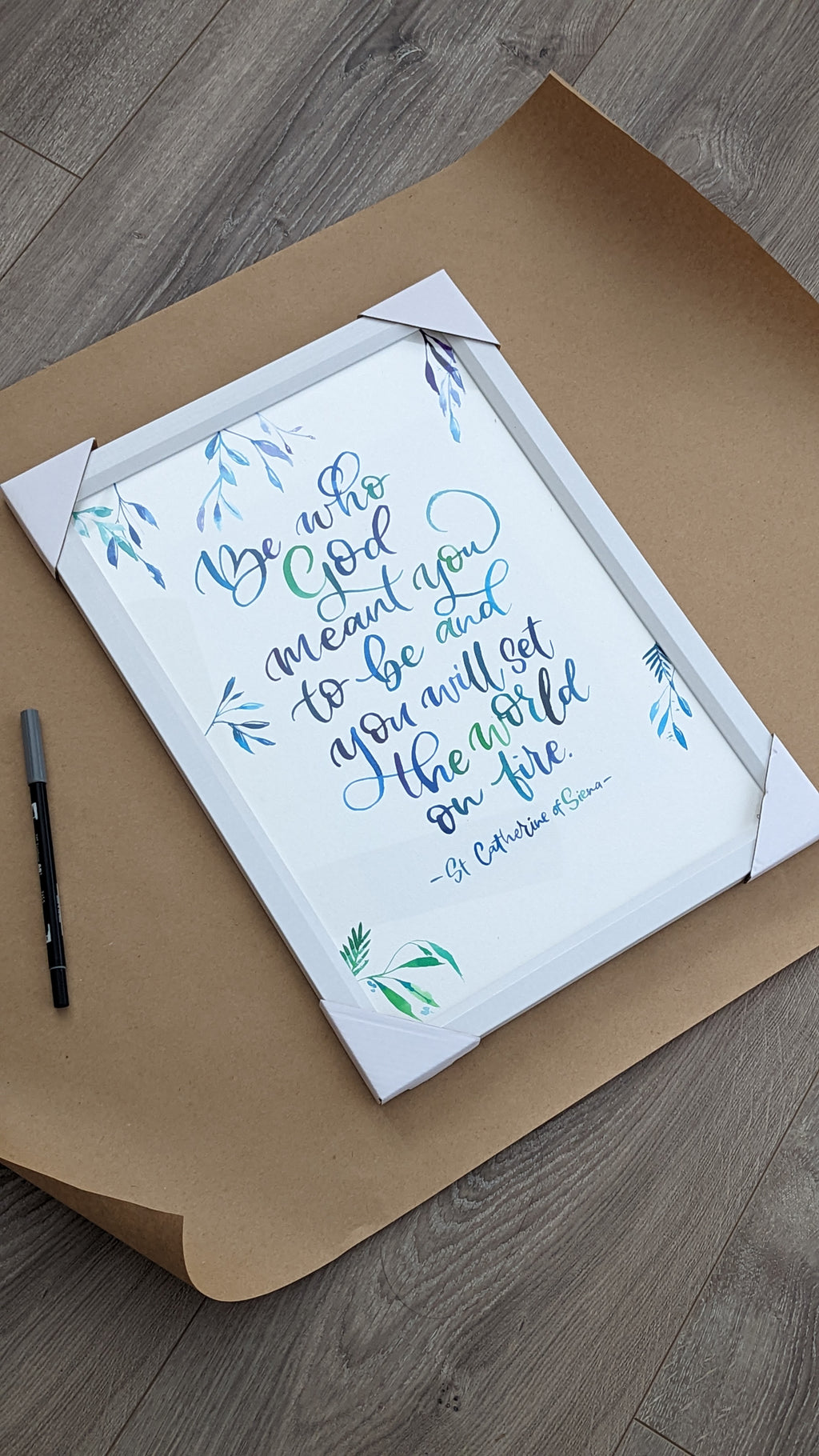 A framed typographic wall art print with a Bible verse in blue and green script, displayed on a brown kraft paper sheet over a gray wood floor. A black marker pen is placed to the left of the frame.