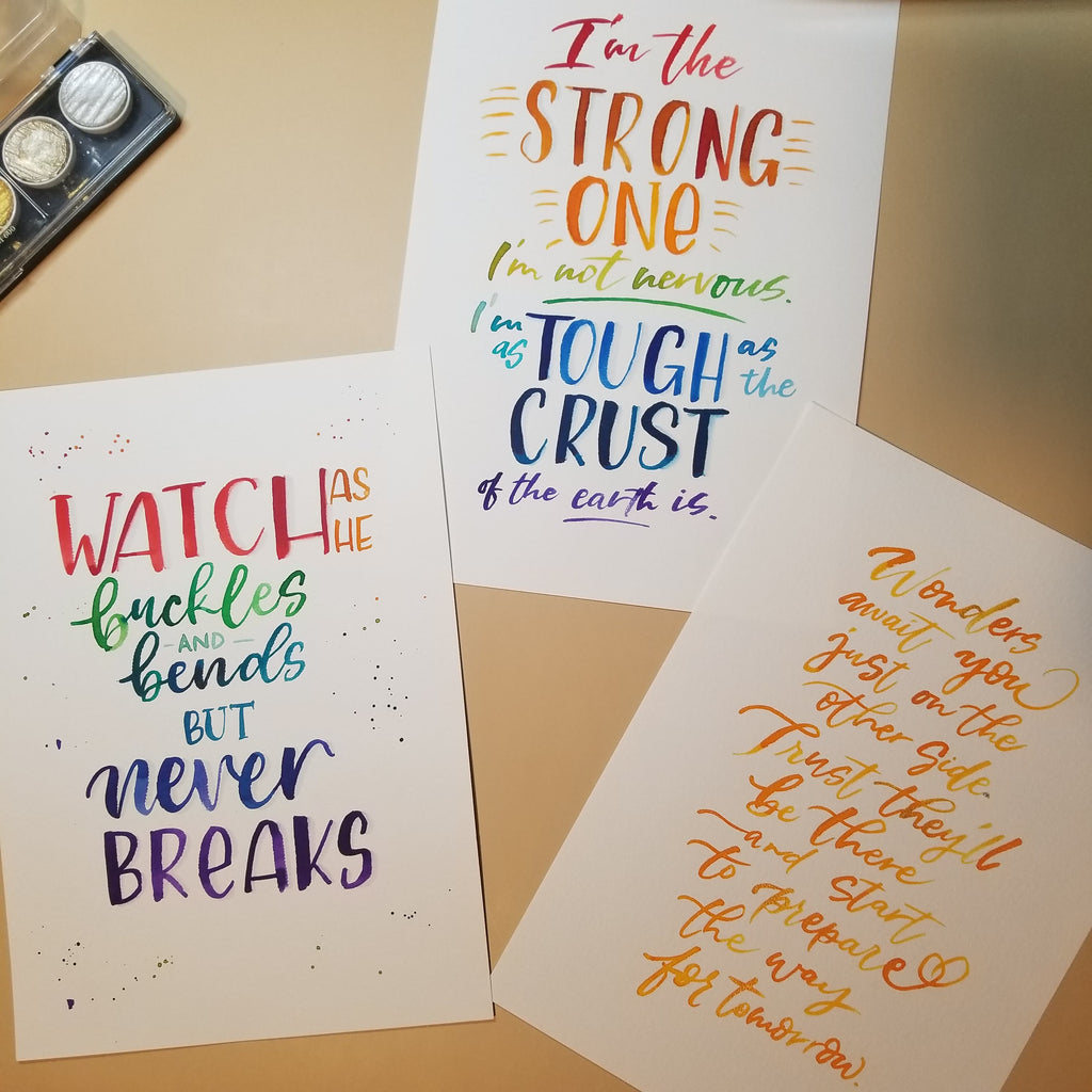Three printed quote cards with colorful hand-lettered text are arranged on a beige surface, alongside a small ink pad. The cards feature inspirational quotes in red, orange, yellow, green, blue, and purple lettering.