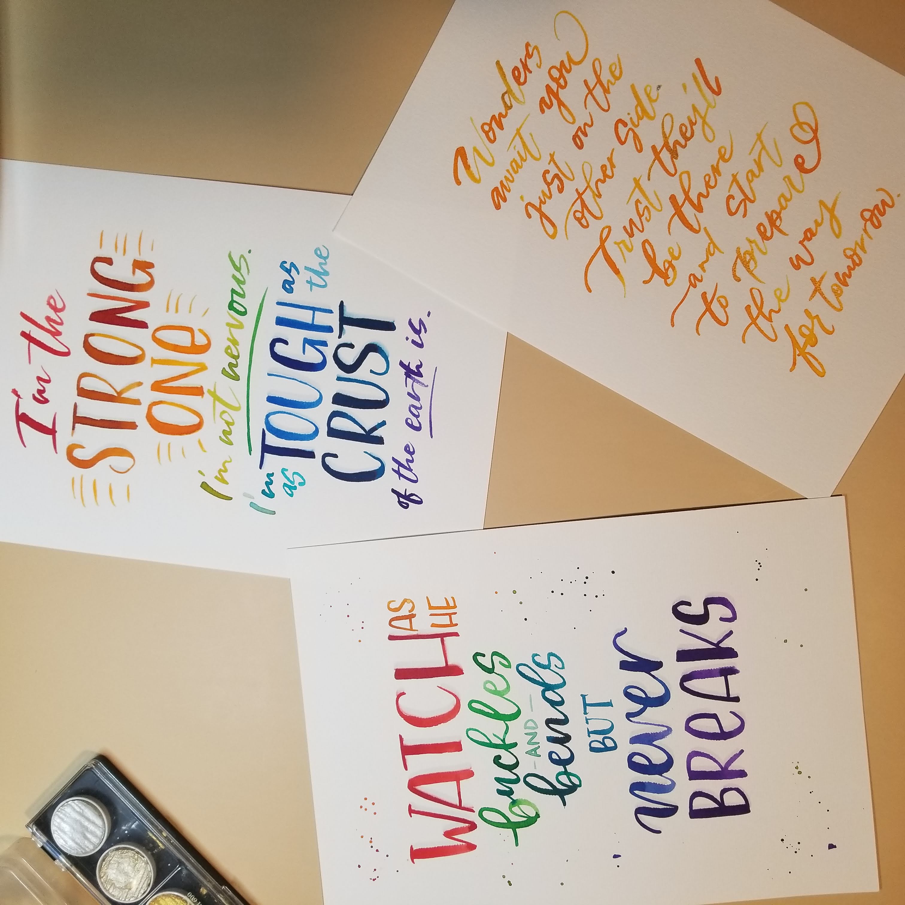 Three printed quote cards with colorful hand-lettered text are arranged on a beige surface, alongside a small ink pad. The cards feature inspirational quotes in red, orange, yellow, green, blue, and purple lettering.