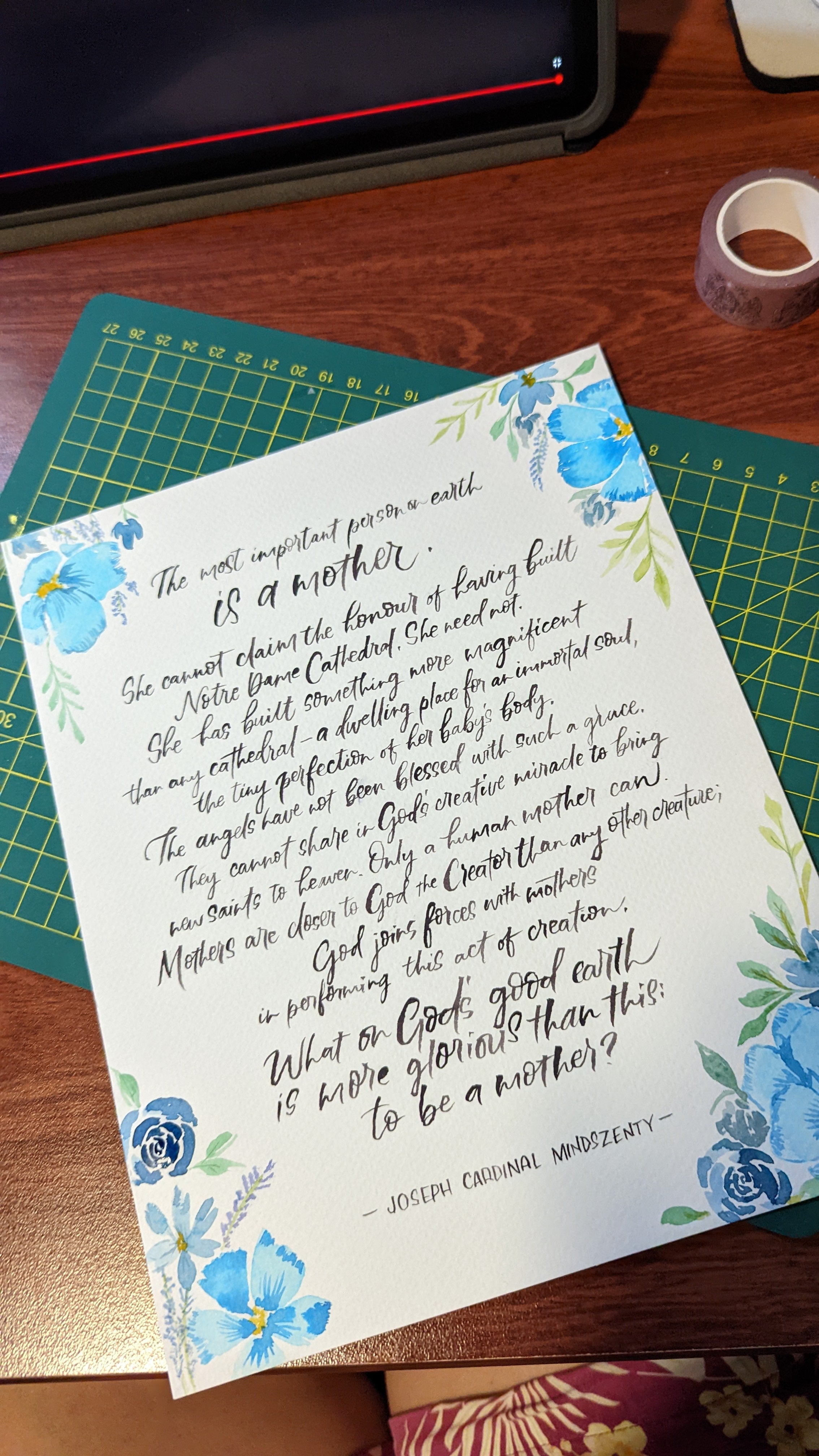 A printed card or letter with cursive text and blue floral illustrations, placed on top of a green cutting mat on a wooden desk. A laptop edge and tape roll are partially visible around it.