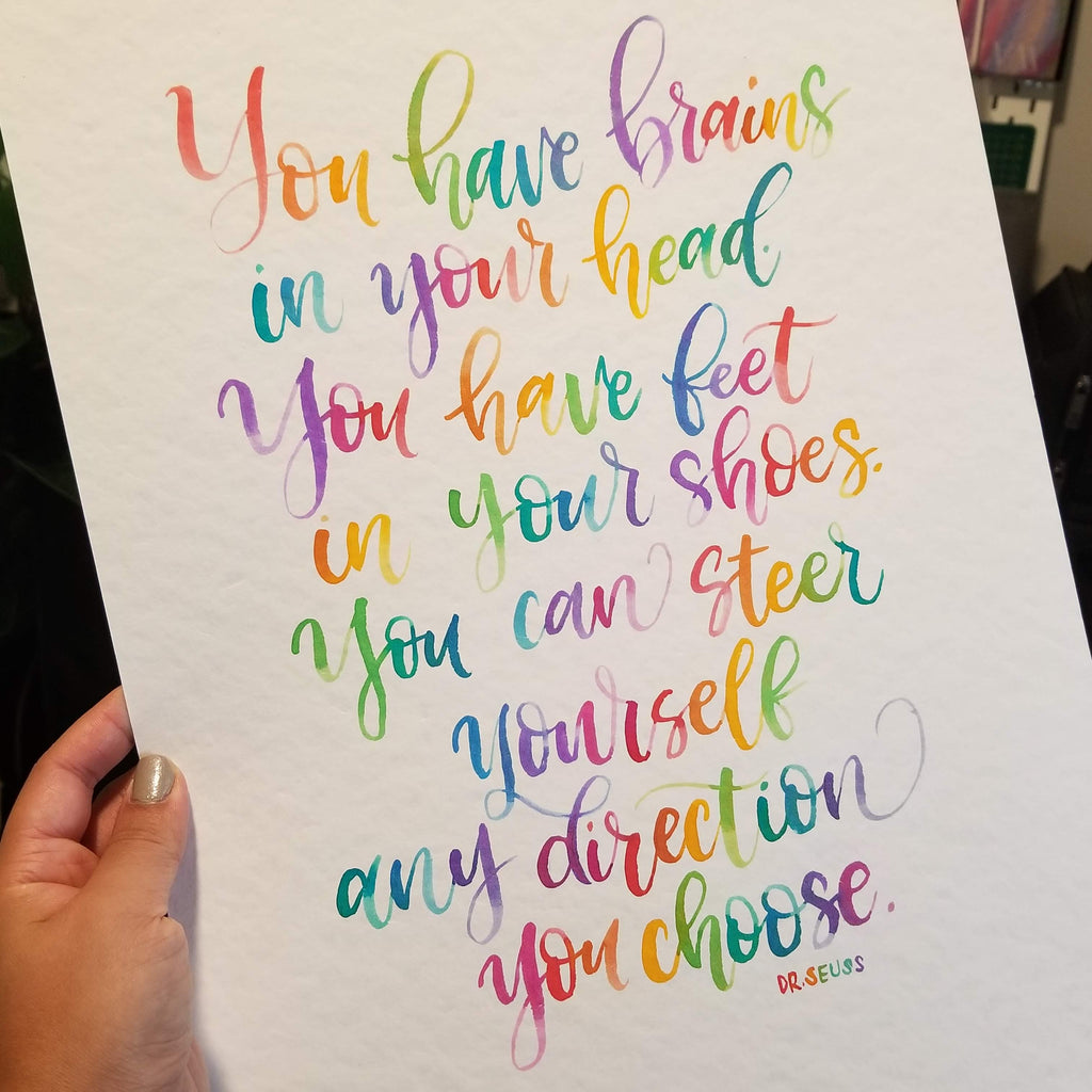 A hand holds a white art print featuring colorful, hand-lettered motivational text by Dr. Seuss.