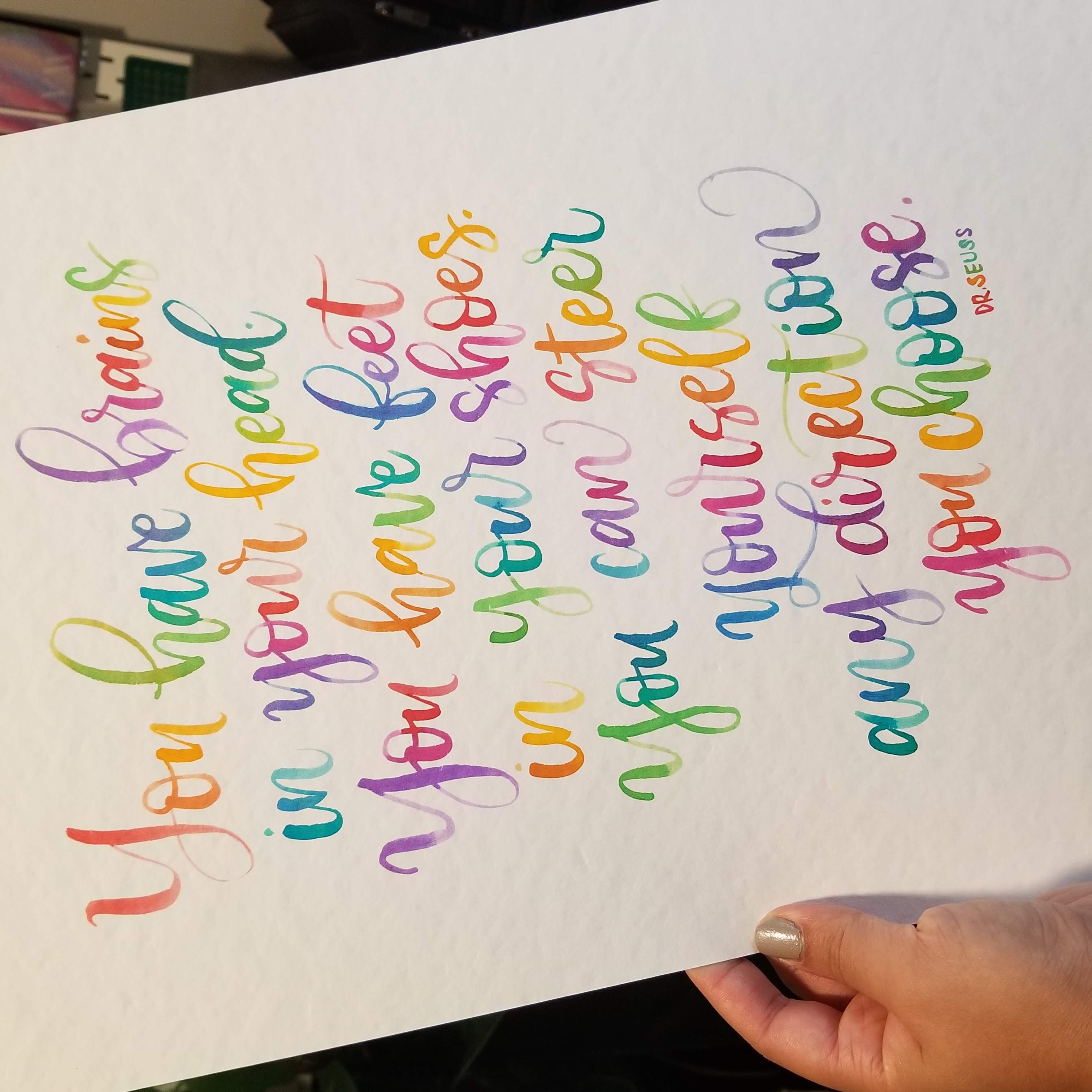 A hand holds a white art print featuring colorful, hand-lettered motivational text by Dr. Seuss.