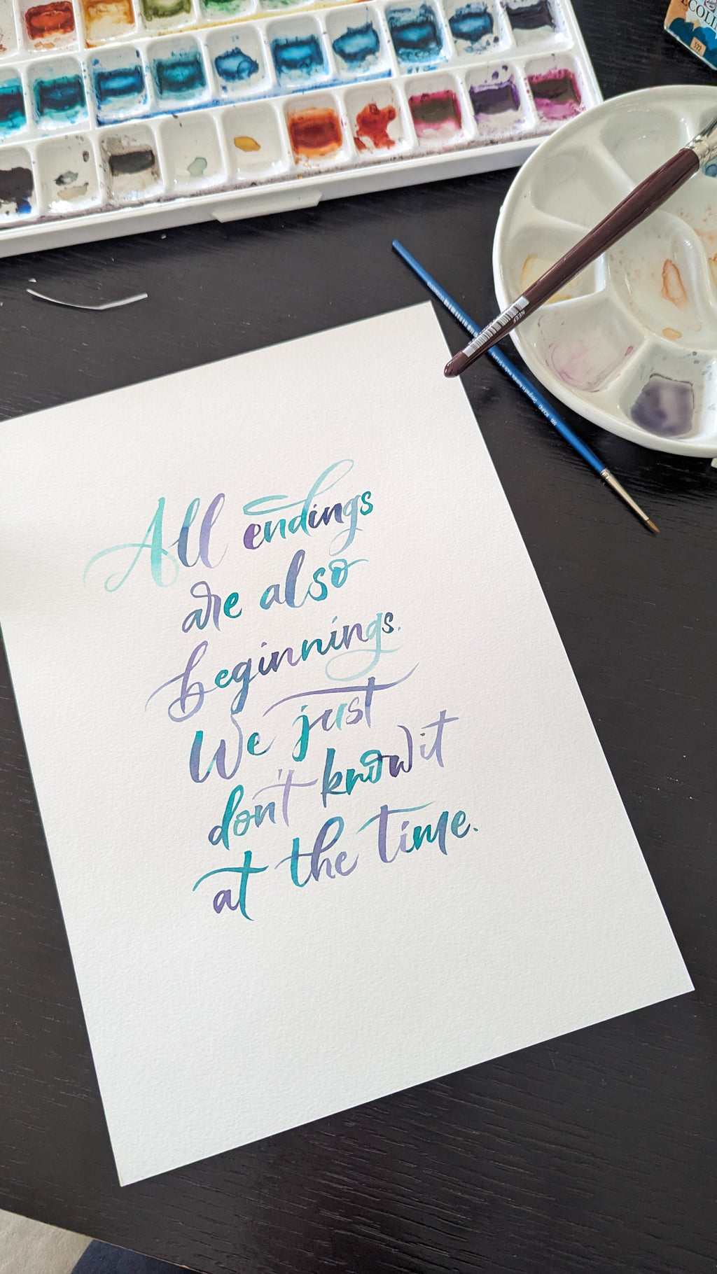 A white sheet of paper with colorful calligraphy text reading 'All endings are also beginnings. We just don't know it at the time.