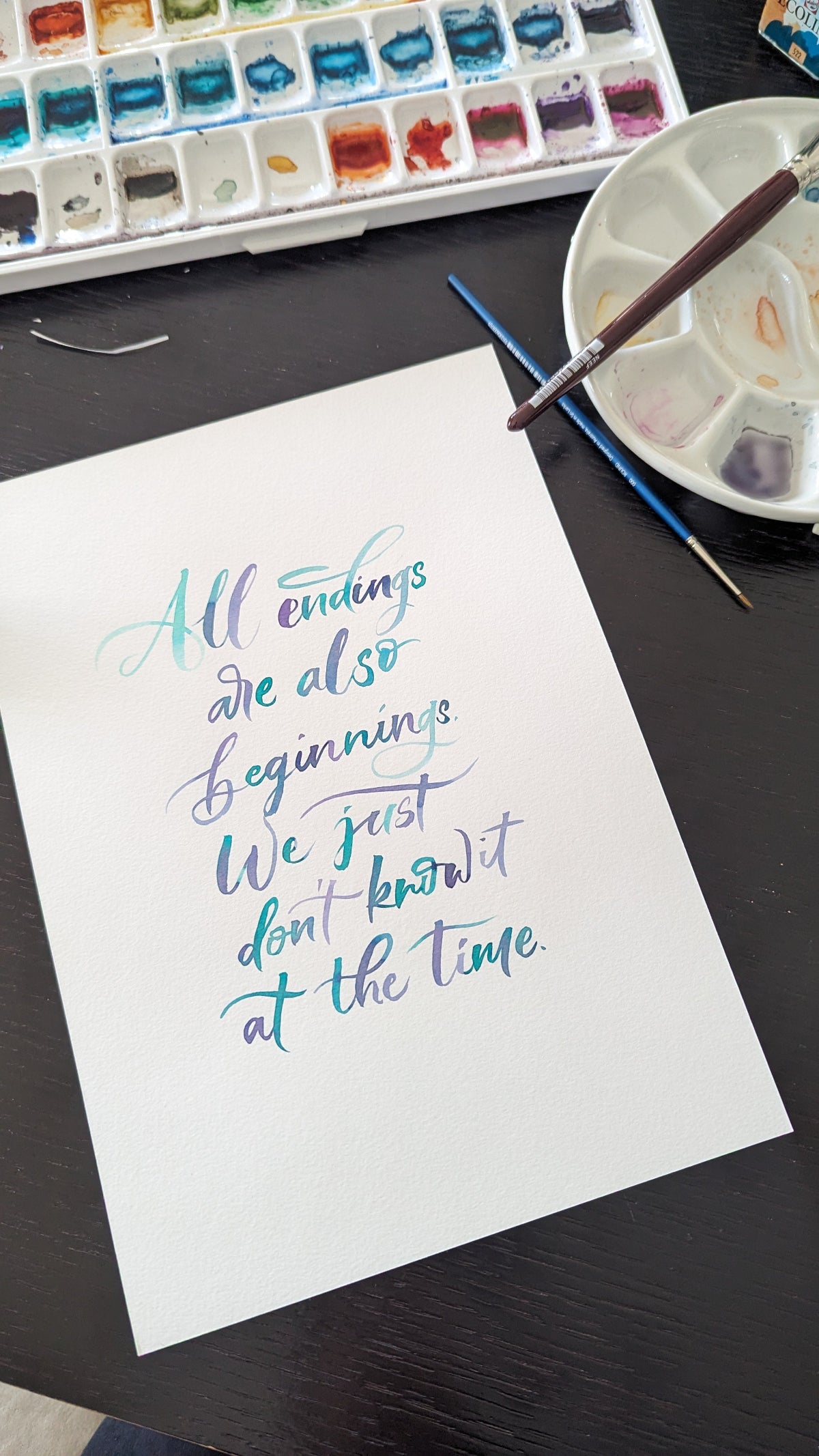 A white sheet of paper with colorful calligraphy text reading 'All endings are also beginnings. We just don't know it at the time.