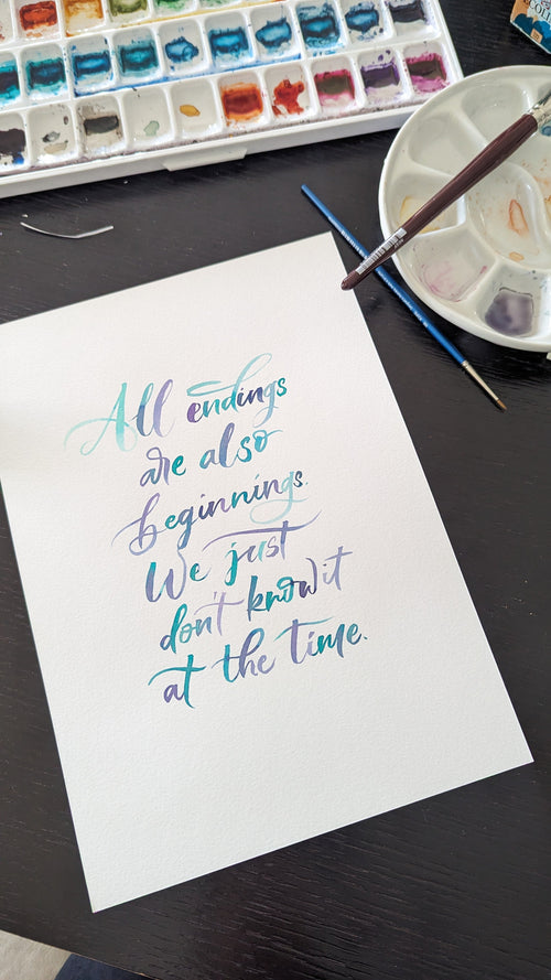 A white sheet of paper with colorful calligraphy text reading 'All endings are also beginnings. We just don't know it at the time.