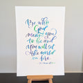 A vertical white art print featuring a colorful hand-lettered Bible verse from Matthew 11:28, displayed on a small wooden easel stand against a neutral light gray wall and dark black tabletop.