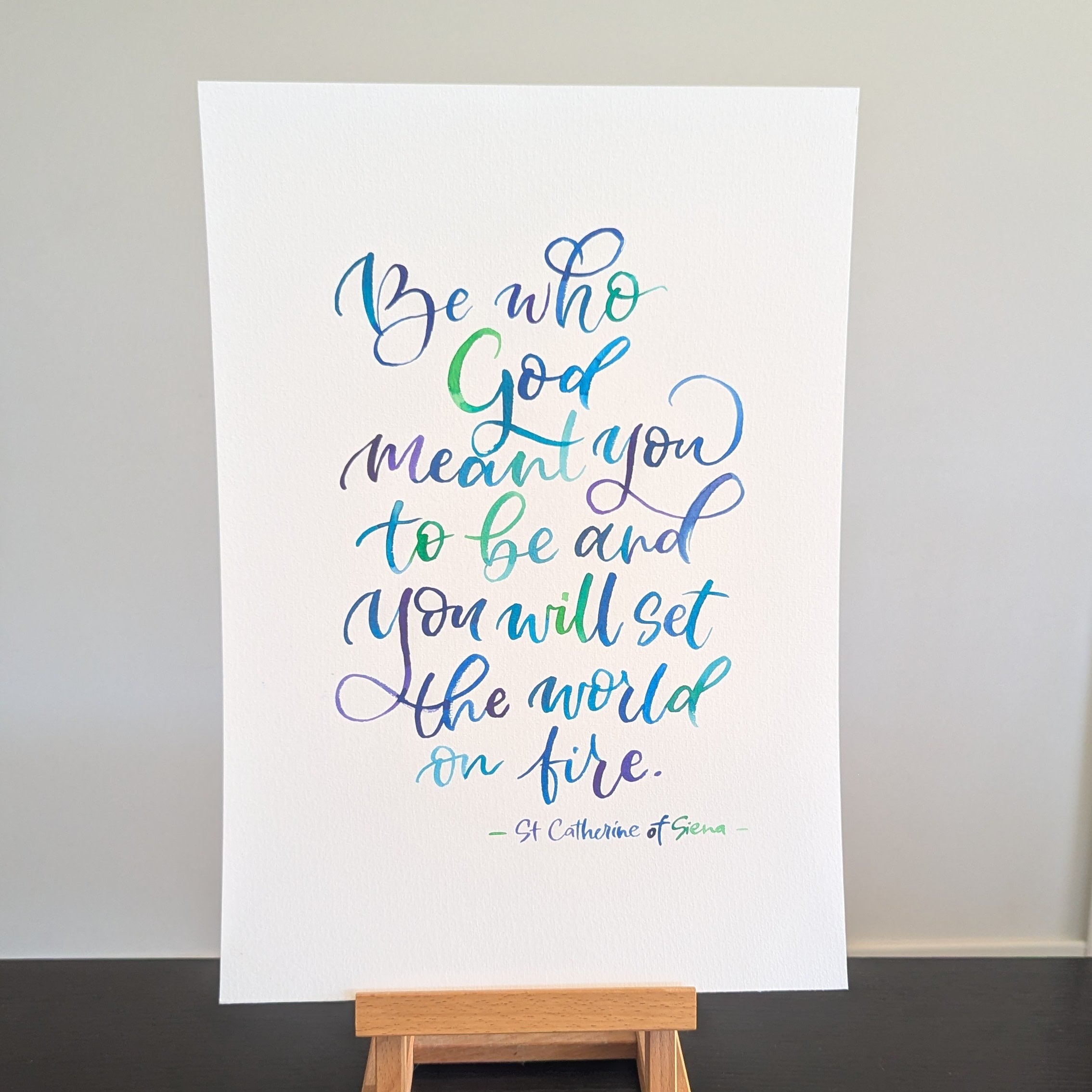 A vertical white art print featuring a colorful hand-lettered Bible verse from Matthew 11:28, displayed on a small wooden easel stand against a neutral light gray wall and dark black tabletop.