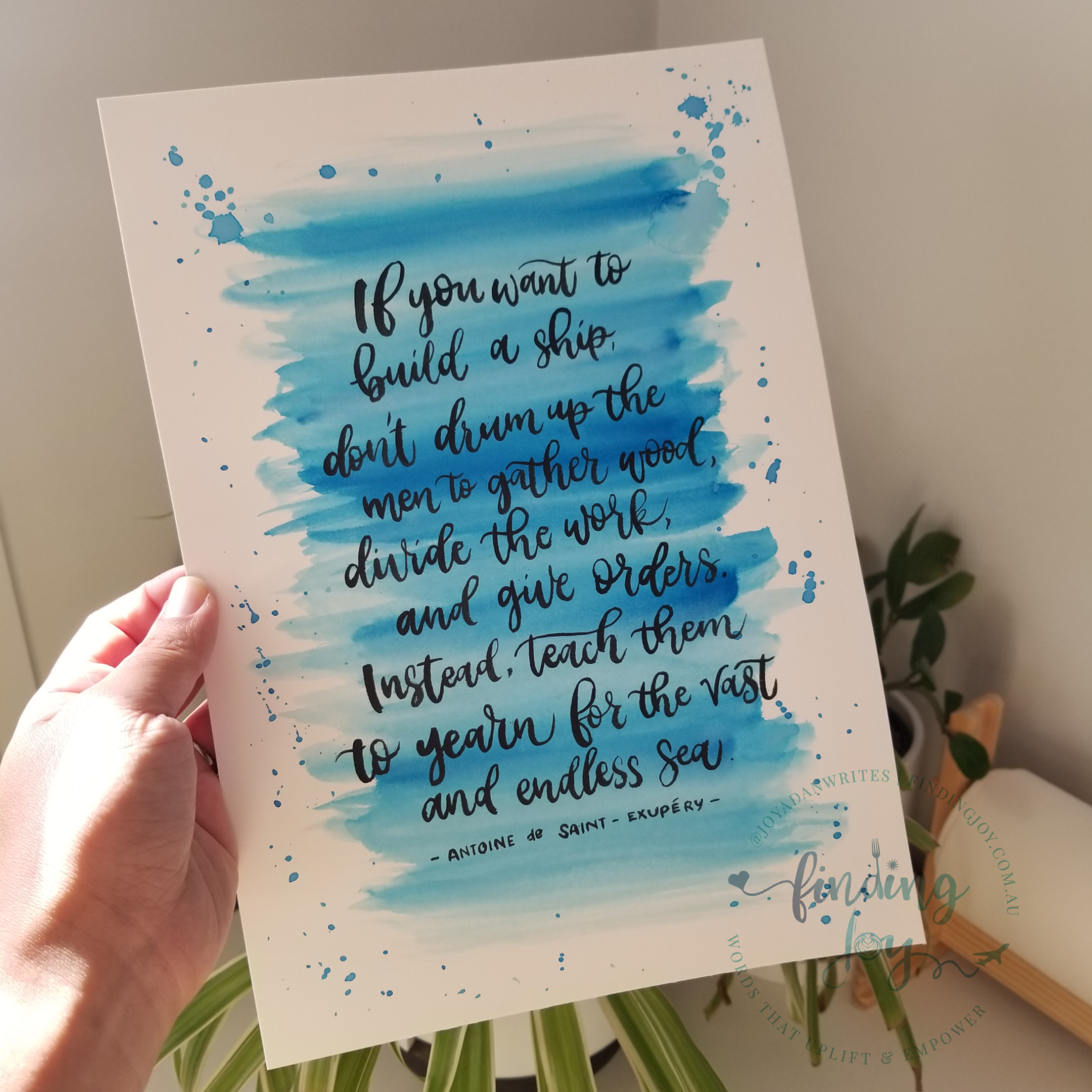 A hand holds a white art print featuring a blue watercolor wash and a handwritten quote by Antoine de Saint-Émery. The print is shown against a softly blurred indoor background with a plant and a wooden object.