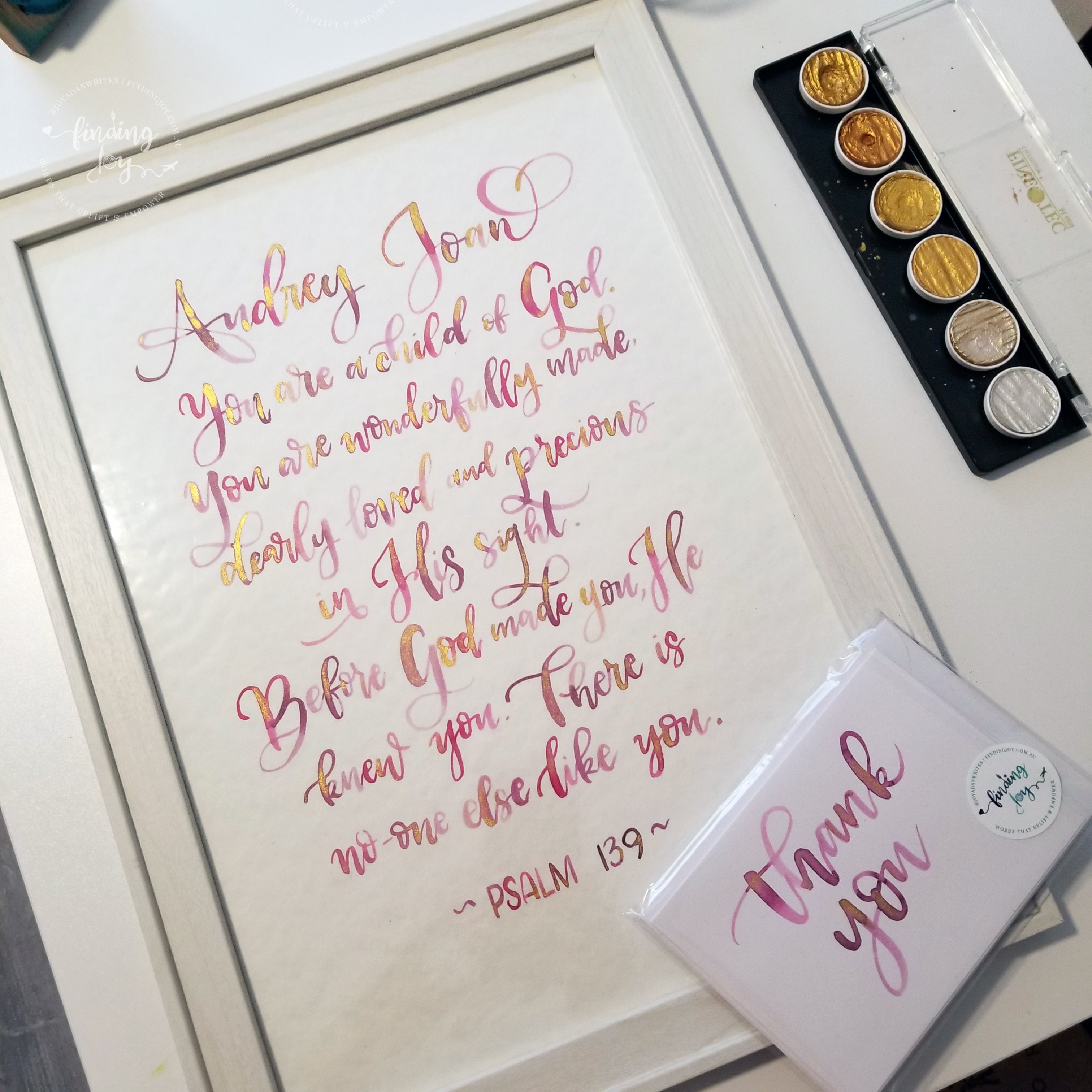 A framed typographic art print featuring a hand-lettered Bible verse in pink and gold metallic script on a white background, styled on a tabletop with a paint palette and a small thank-you card.