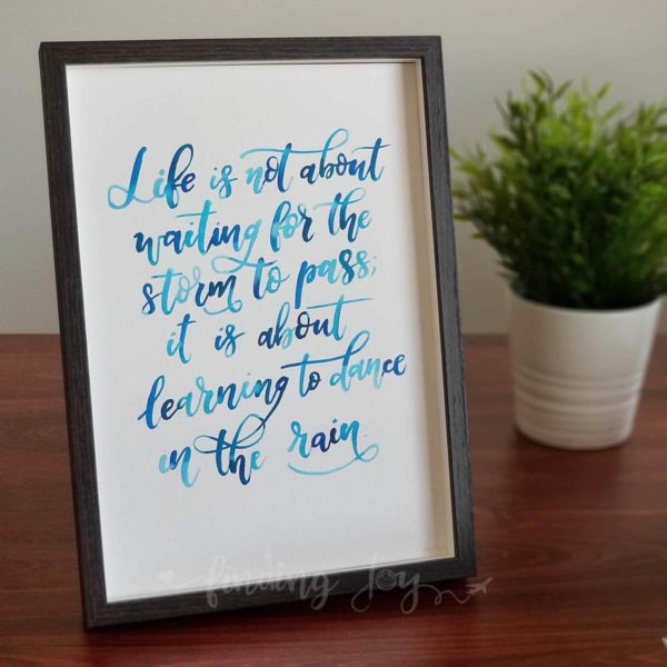 Original, hand-painted brush-lettered artwork of the quote "Life is not about waiting for the storm to pass, it is about learning to dance in the rain" written in different types of blue watercolour on A4 300GSM card. © Joy Adan 2018