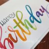 Hand-painted greeting card with 'Happy birthday' hand-lettered in multicolour/rainbow Ecoline ink. Each card is lettered on matte card stock. Inside: blank. © Joy Adan 2018