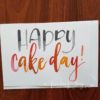 "Happy Cake Day" Greeting Card written in watercolour brush calligraphy in black, orange and red © Joy Adan