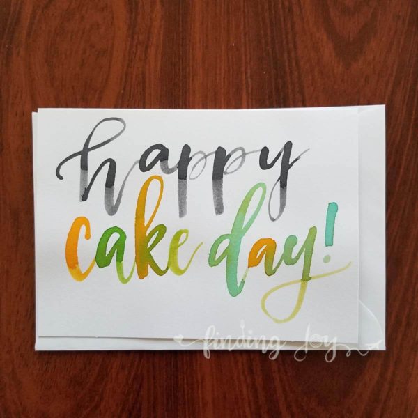 "Happy Cake Day" Greeting Card written in watercolour brush calligraphy in black, yellow and green © Joy Adan
