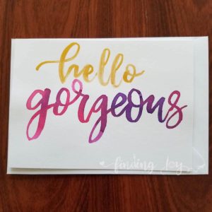 Greeting card with "hello gorgeous" hand-lettered in gold and fuschia Ecoline ink. Each card is painted on matte card stock. Inside: blank. © Joy Adan 2018