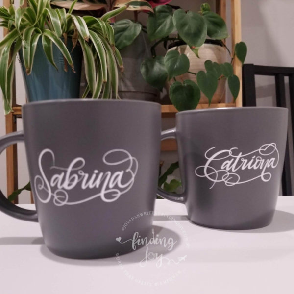 2 personalised grey mugs. One with "Sabrina" in white calligraphy and the other with "Catriona" in white calligraphy.